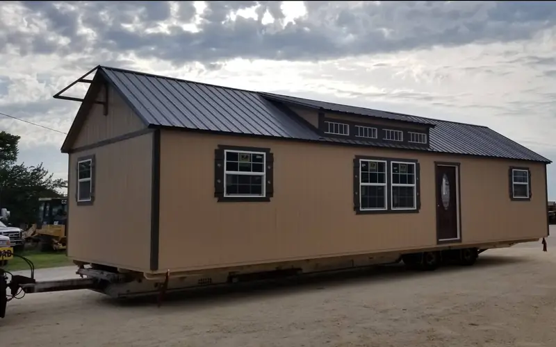A tan cabin with dormer windows hitched on a delivery trailer